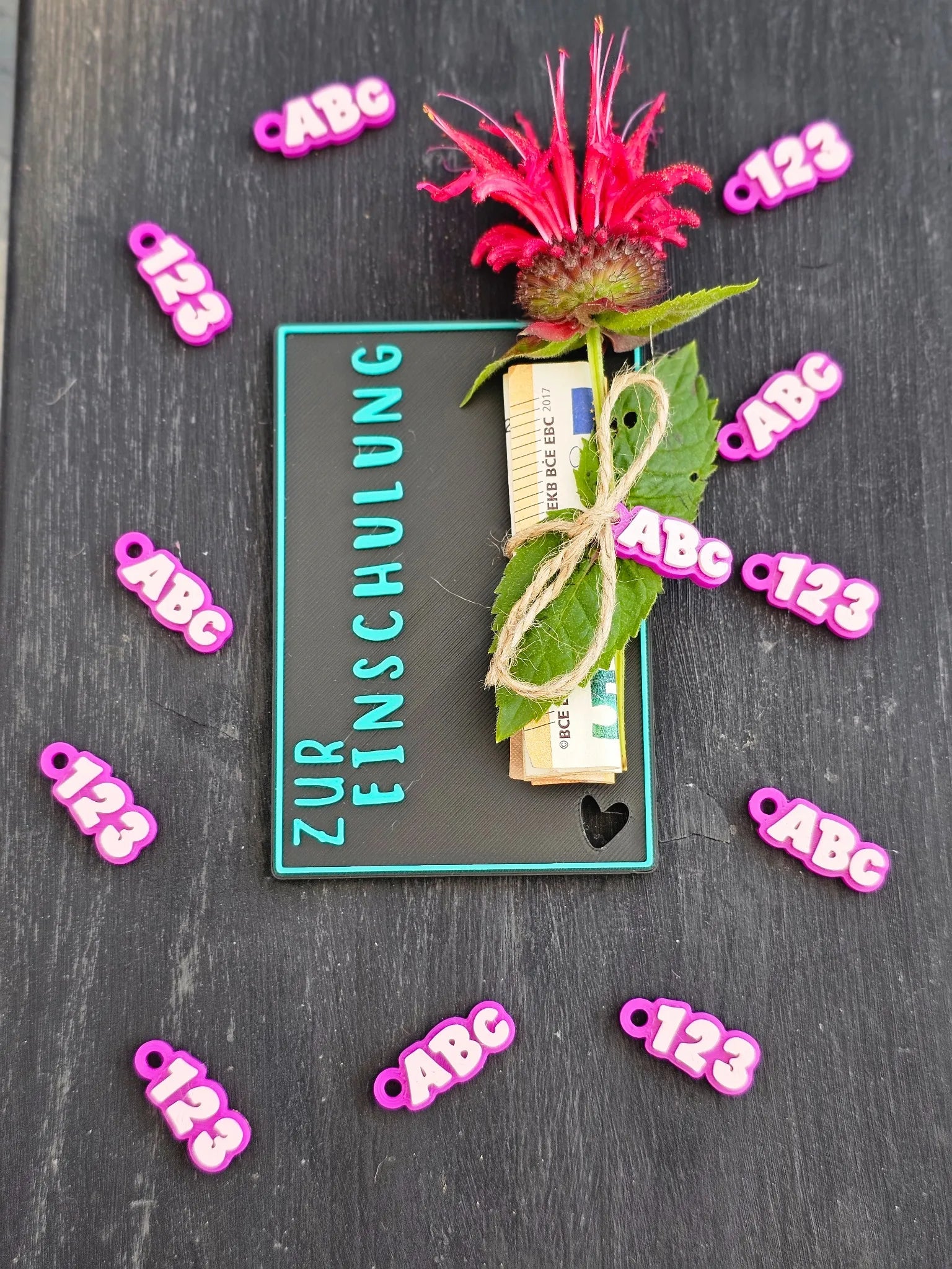 Schwarze WunschWerk-Karte mit türkisfarbener Aufschrift „Zur Einschulung“, Blume, Geldschein und bunter ABC/123-Deko
