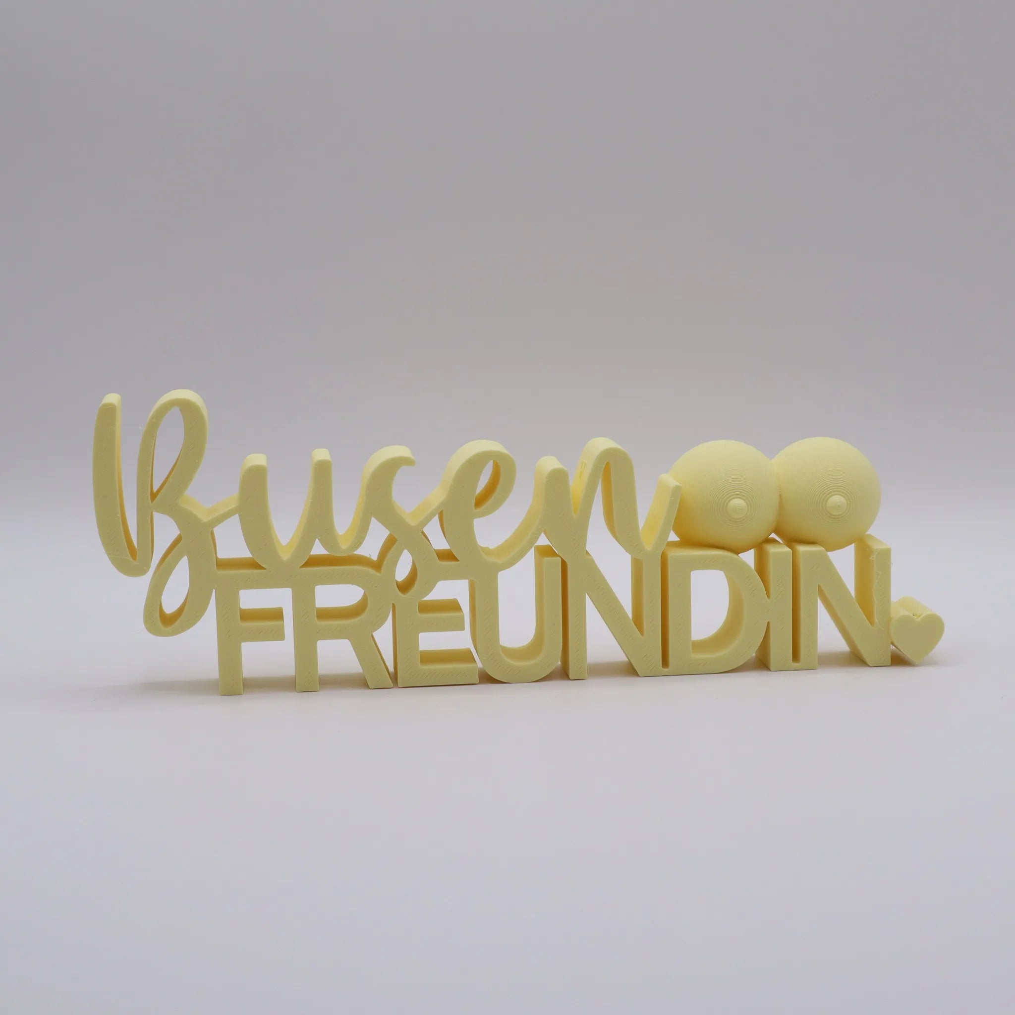 3D-gedrucktes Deko-Statement in Mandelgelb mit dem Schriftzug Busenfreundin, verziert mit Busen-Symbolen und Herz