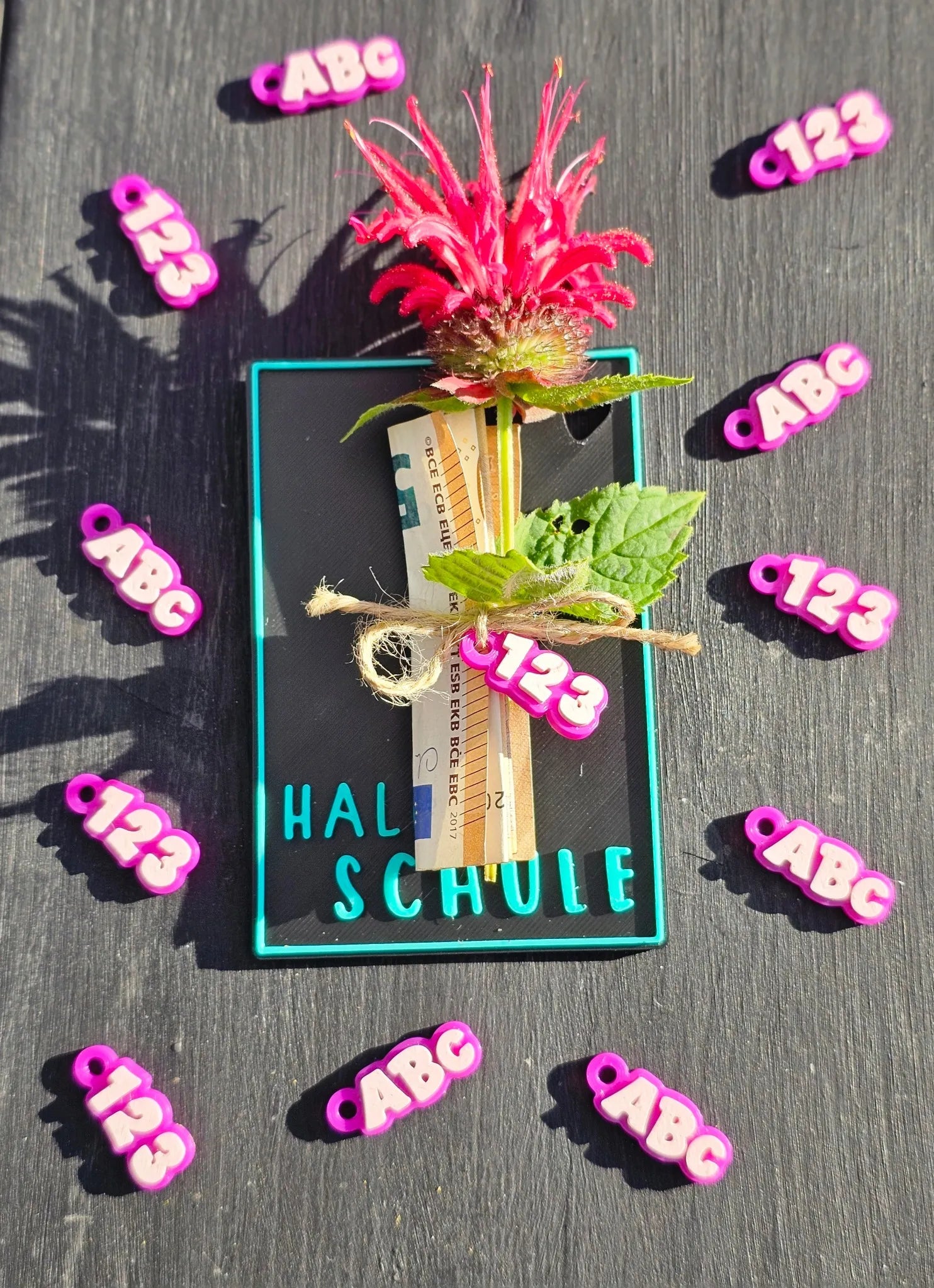 Einschulungskarte mit Blume, Geldrolle und türkisfarbenem Text „Hallo Schule“ auf schwarzem Hintergrund, umgeben von bunten ABC-Elementen

