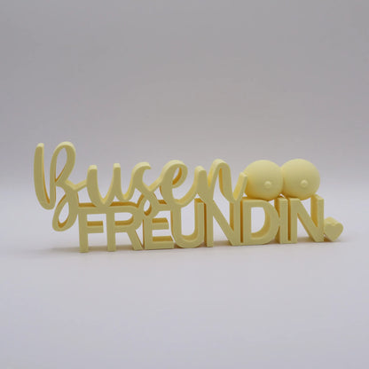 3D-gedrucktes Deko-Statement in Mandelgelb mit dem Schriftzug Busenfreundin, verziert mit Busen-Symbolen und Herz